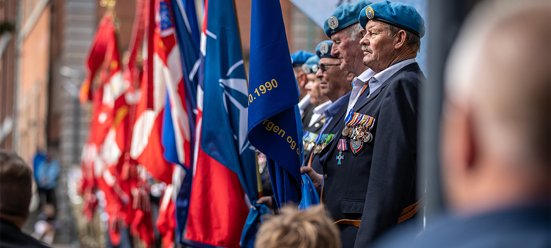 På en lige række står faner med Dannebrog og Nato våbenskjold i en parade. Man kan se, at de faner tættest på kameraet bliver holdt af ældre mænd med blå baretter og mørke habitjakker med anlagte medaljer.