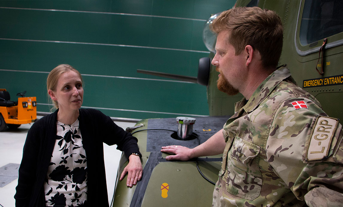 Kvinde i skjorte og blazer taler med en mand i uniform i en stor hangarbygning. De hviler begge armen på vingerne af en helikopter.