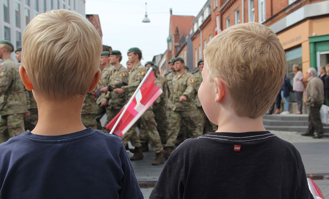 To drenge i forgrunden af billedet med flag i hånden kigger på parade af uniformerede soldater, der går forbi i bymiljøet.