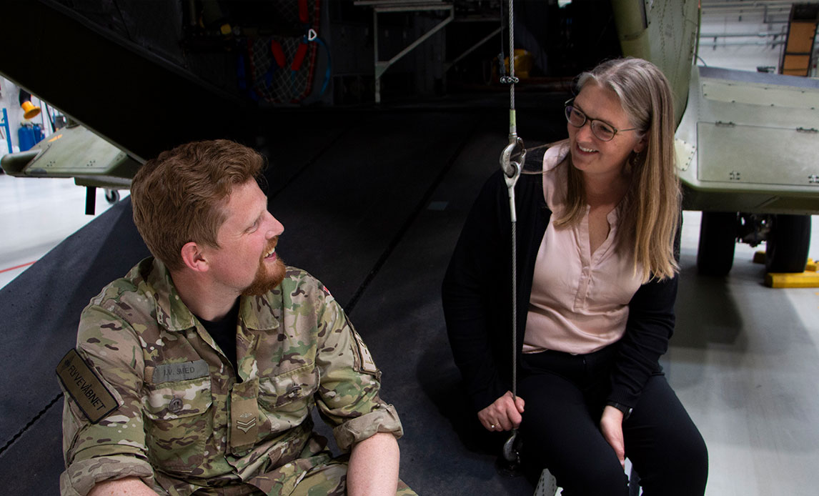 Mand i uniform og en kvinde i civil sidder på klappen af en åben helikopter og smiler til hinanden.