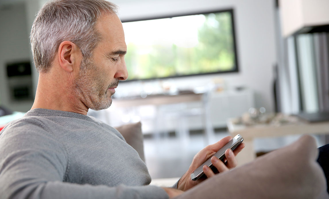 Mand med gråt kort hår og kort fuldskæg sidder i sofa i lys stue med sin smartphone.