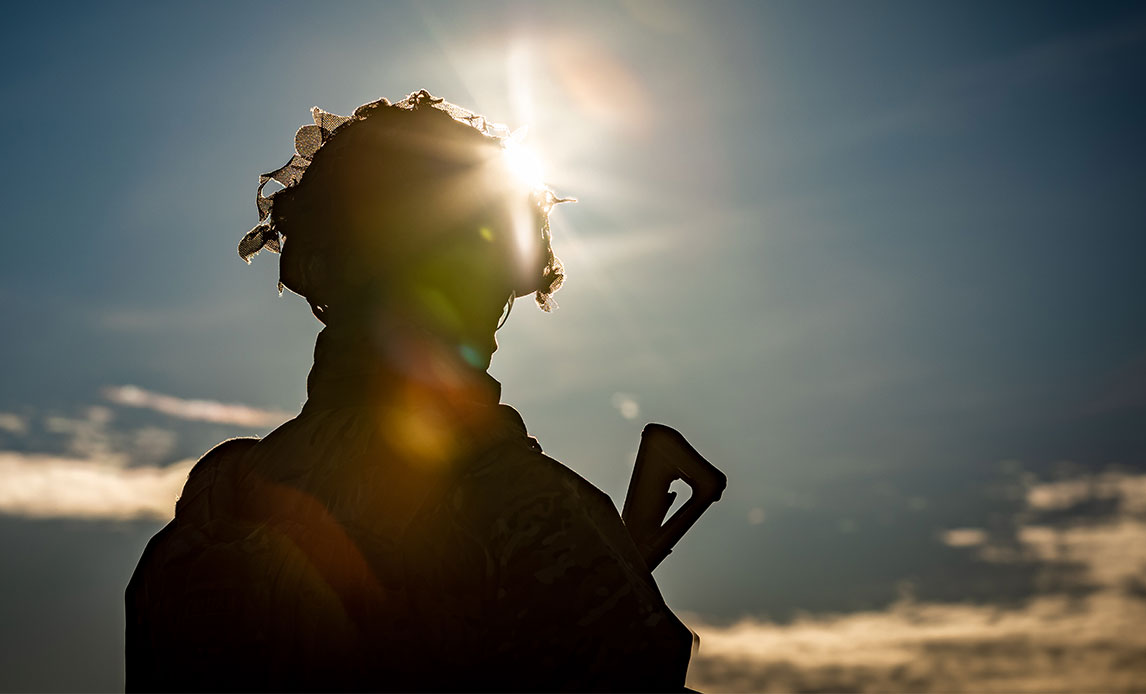 En soldat i uniform står i modlyset. Solen rammer soldatens hjelm, så manden eller kvinden kun fremstår som en skygge. Vedkommende har hævet bunden af sit gevær til over skulderhøjde.