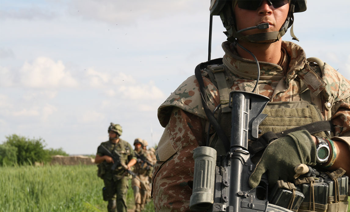 Soldat med hjelm og solbriller, hvor billedet er skåret lige under hjelmkanten. Han går med gevær gennem grønt område.
