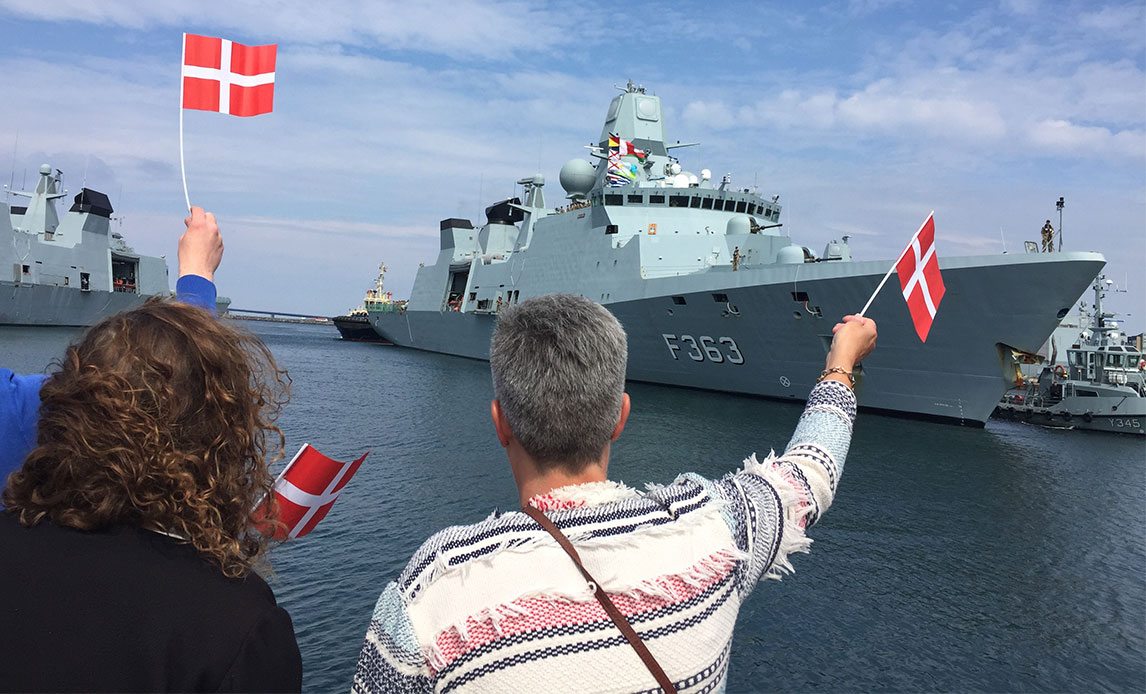 Karseklippet kvinde står i striktrøje på havnekaj og vifter med et plastikflag mod gråt skib, korvetten Niels Juel. Ved siden af hende står en kvinde med skulderlagt krøllet hår også og vinket med et flag.