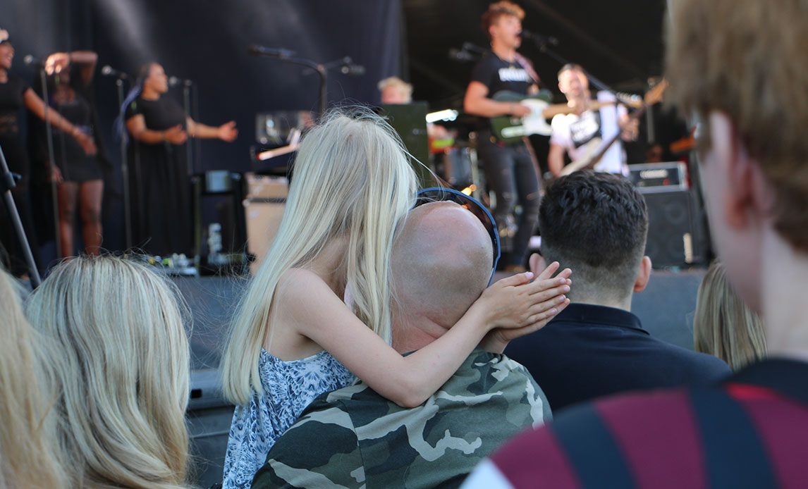 Mand i camouflagefarvet t-shirt bærer på blond og langhåret pige blandt andre koncertpublikummer. Foran dem ses bandet med forsanger i front.