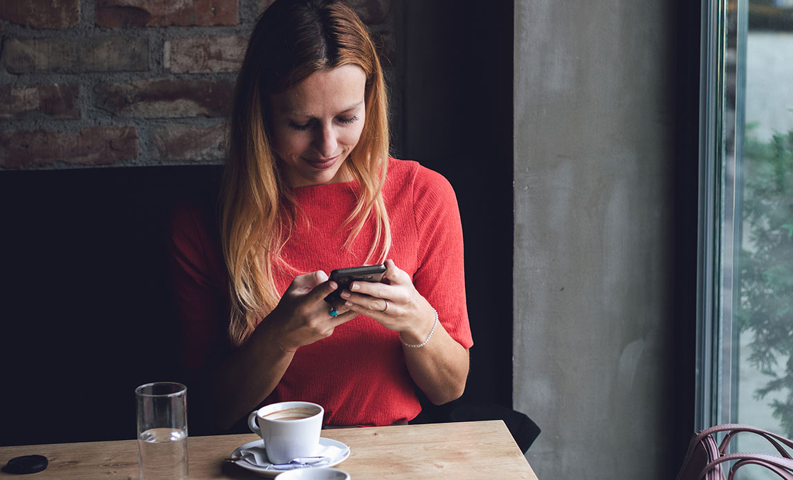 Kvinde sidder med smartphone i hånden foran et cafébord med en kop kaffe.