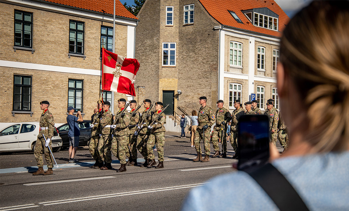 En række af soldater marcherer gennem et ældre bybillede med sandfarvede bygninger. Forrest går en soldat med en fane. I forgrunden af billedet står en kvinde og tager foto af de marcherende soldater med sin telefon.