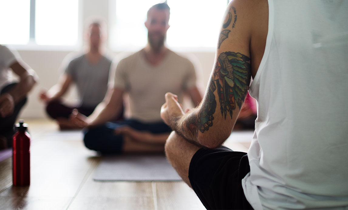 Mænd sidder i skrædderstilling med hænderne hvilende på knæene i en gymnastiksal. Manden tættest på kameraet er i korte bukser og ærmeløs t-shirt og har tatoveringer på overarm og skulder.
