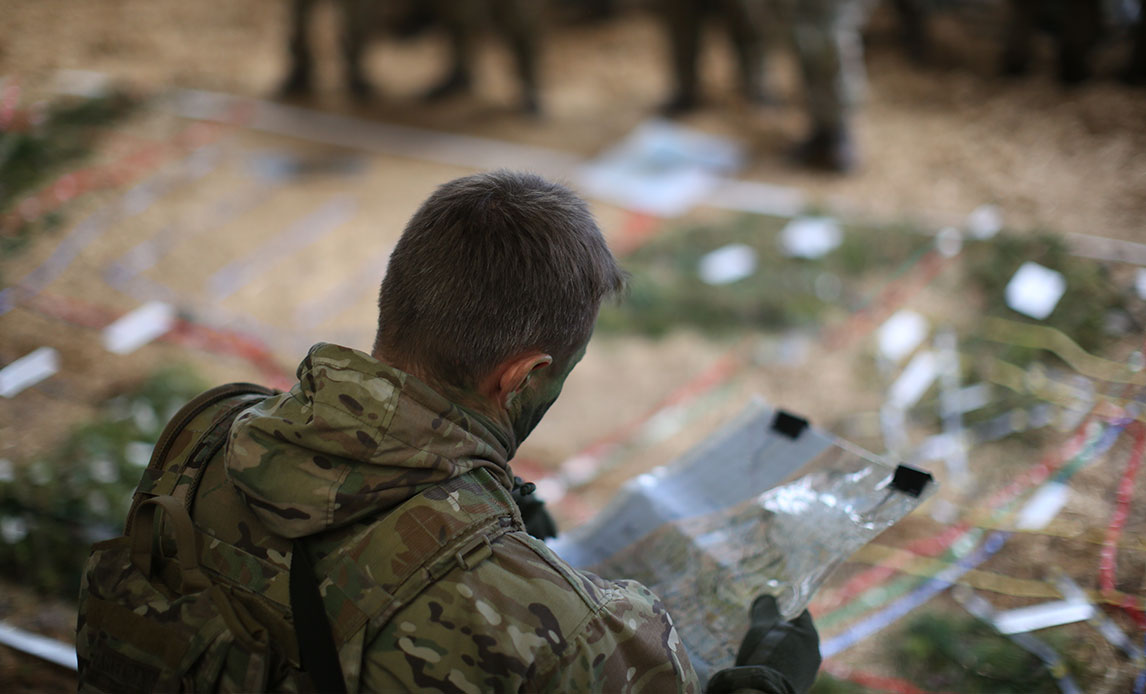 Uniformeret soldat med kort hår og sløring i ansigtet holder et lamineret landkort i hænderne og studerer det. Soldaten står tilsyneladende i en lade med halm på gulvet.