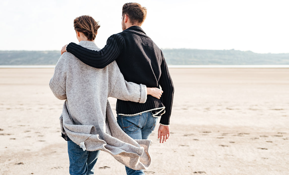 Kvinde og mand på stranden i blæsevejr. Hendes korte hår og cardigan kastes bagud af vinden. Han holder en arm rundt om hendes skulder