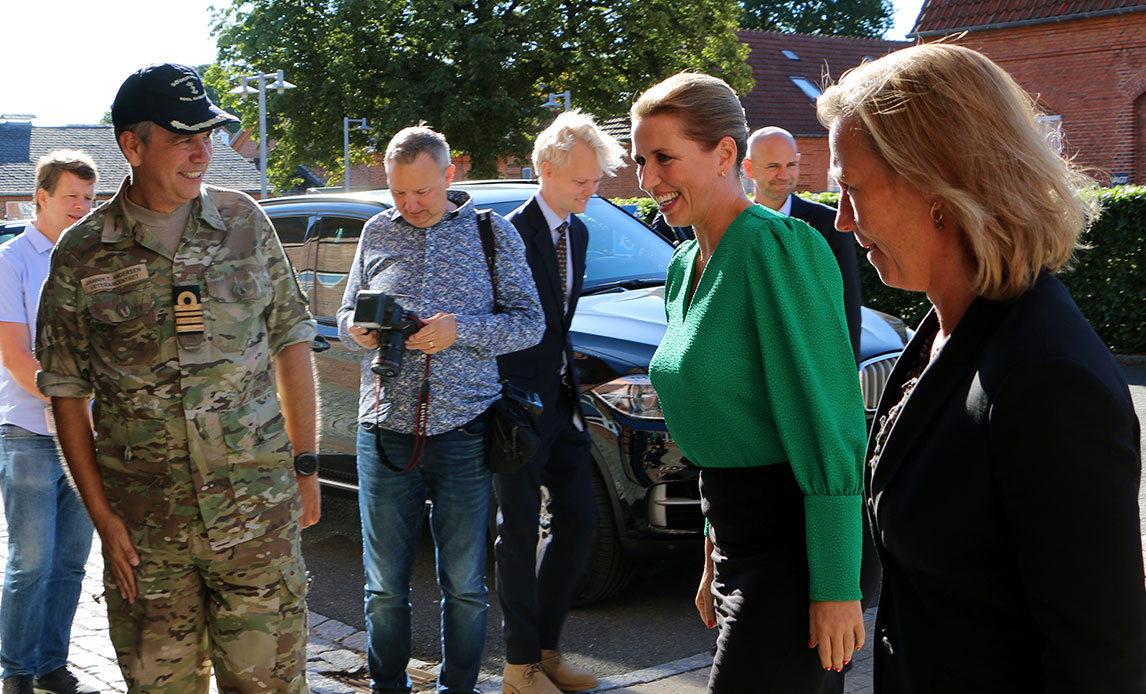 Statsminister Mette Frederiksen træder nærmere en uniformeret mand med kasket, der slår ud med hånden for at få hende til at træde nærmere. Rundt om dem en mindre mængde af mennesker.