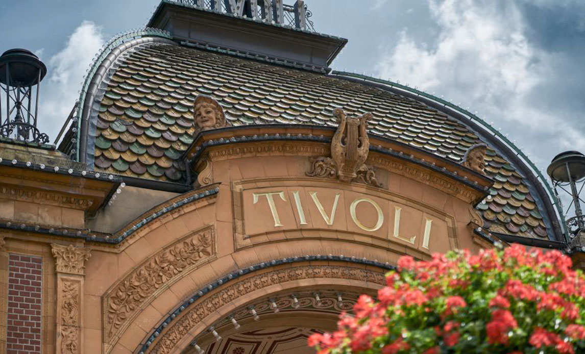 I frøperspektiv et zoomet billede af en buet port af terracottafarvede mursten, hvor der med gyldne bogstaver står Tivoli.  Bag buen en blå himmel og i forgrunden en masse blomster fra en ophængt blomsterkrukke.