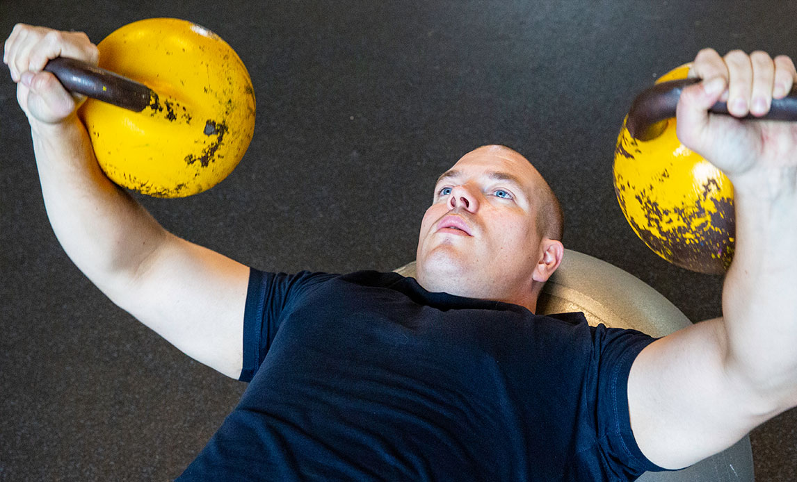 Mand ligger med skuldrene på, hvad der ligner en yogabold eller mindre pude. Hver af hans hænder løfter to gule, men slidte kettlebells op over hans hovedet.