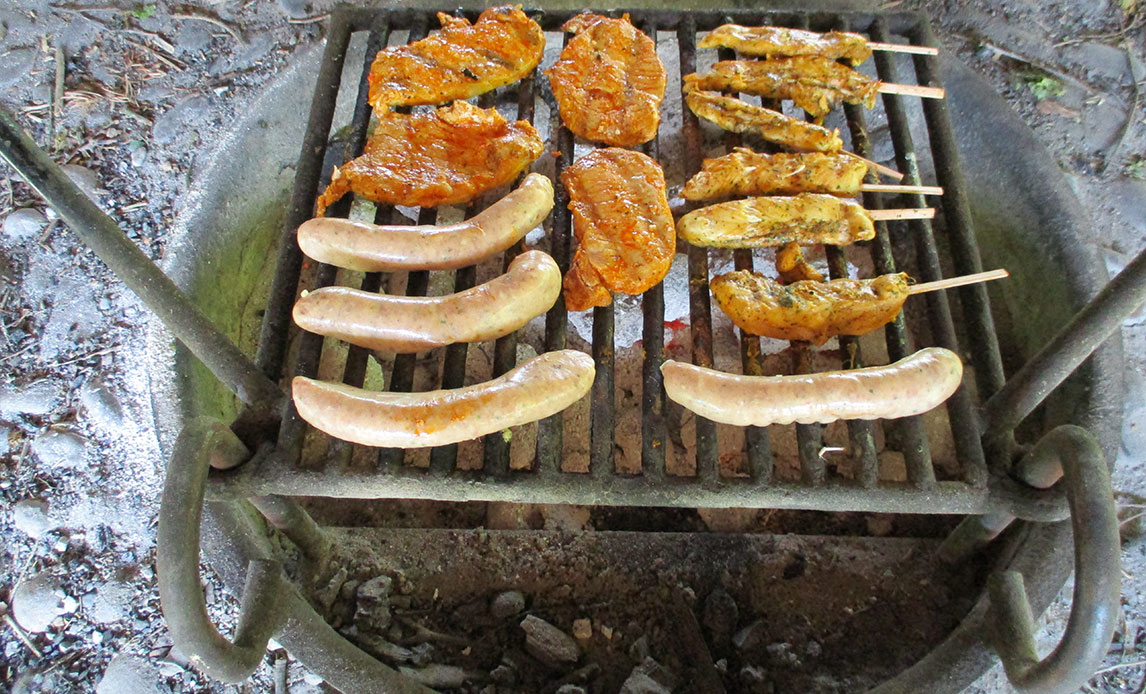 Nærbillede af tændt bål med grillrist med marinerede kyllingefileter, kyllingespyd og pølser.