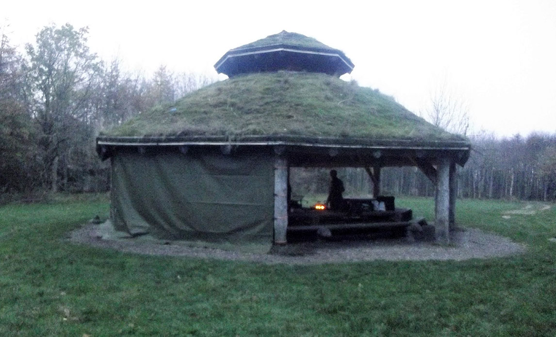 Bålhytte i tusmørke med grønne arealer omkring. Der er monteret en presenning på bålhyttens ene side. Inde i hytten anes et bålsted i mørket.