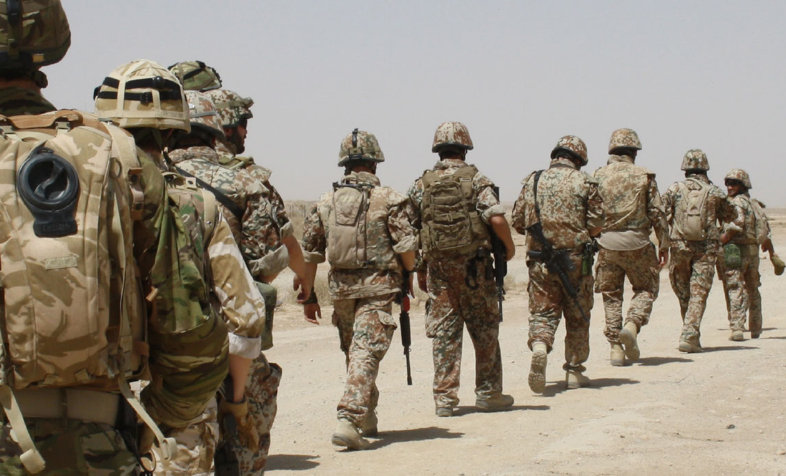 En række af soldater i sandfarvet uniform går i ørkenlignende terræn med ryggen til. Nogle bærer rygsække, andre camelbacks med vand på ryggen, men de bærer alle hjelm.