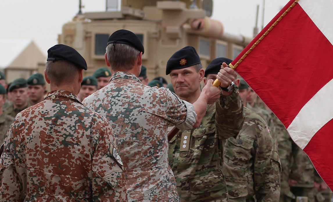 To uniformerede mænd står overfor hinanden, mens den ene rækker en rød og hvid fane til den anden. Bag dem står soldater opstillet.