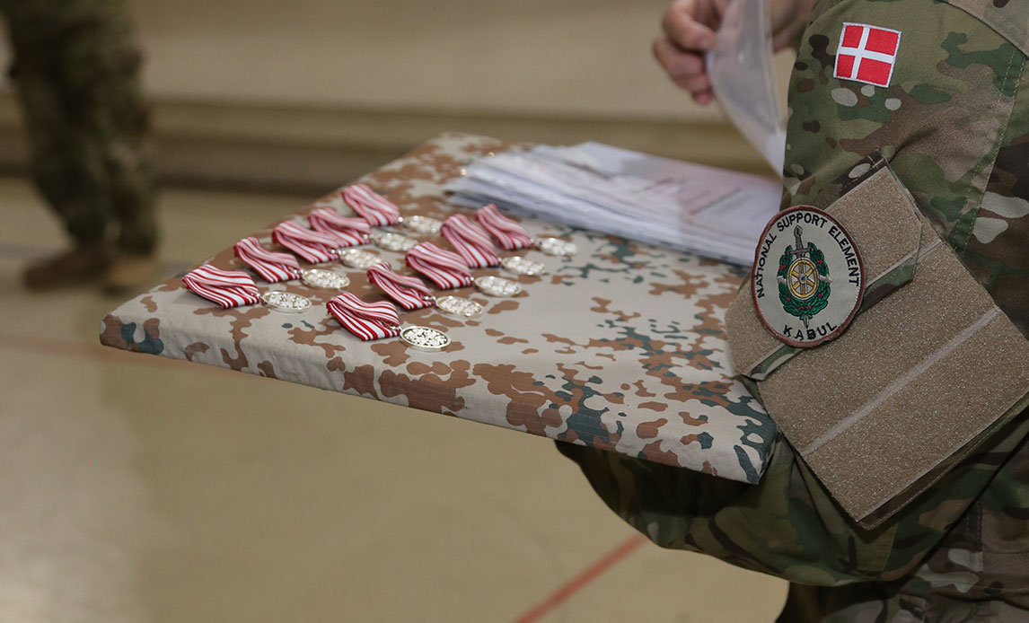 Arme på en uniformeret soldat holder en plade, hvor der på det sløringsfarvede stof henover pladen ligger et antal medaljer klar til at blive uddelt.