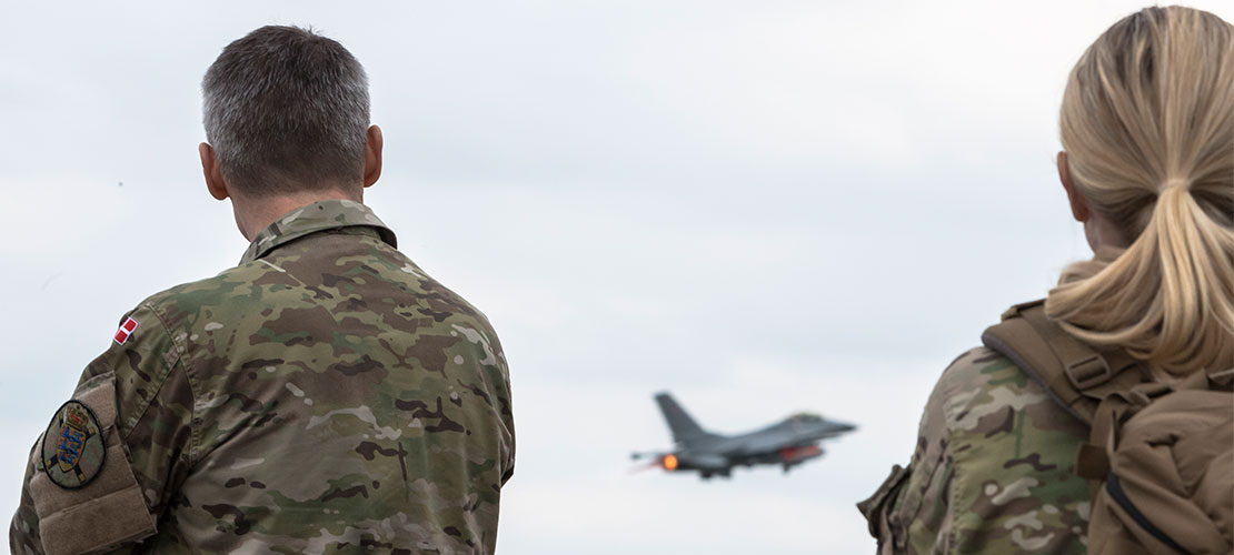 En korthåret uniformeret mand står i venstre side af billedet, mens en langhåret, uniformeret kvinde står i højre side af billedet. Begge kigger de på en F16, der flyver på distancen.