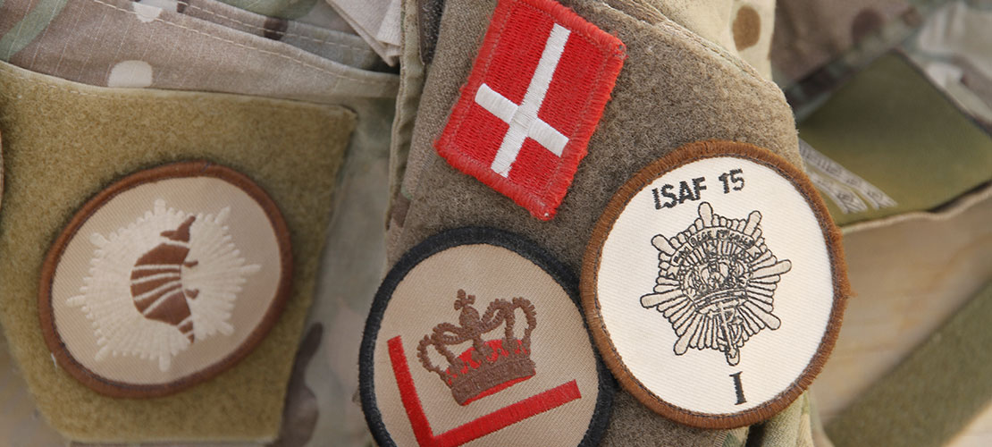 Nærbillede af uniforms ærmer, hvor et lille Dannebrog, et mærke med ISAF hold 15, et mærke med en krone og et mærke med et bæltedyr sidder fast på velcrostykket.