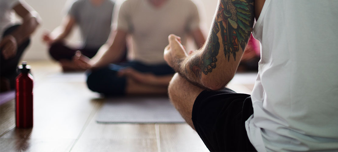 Mand med stor tatovering på venstre overarm sidder i undertrøje og shorts i skrædderstilling foran andre personer på yogamåtter