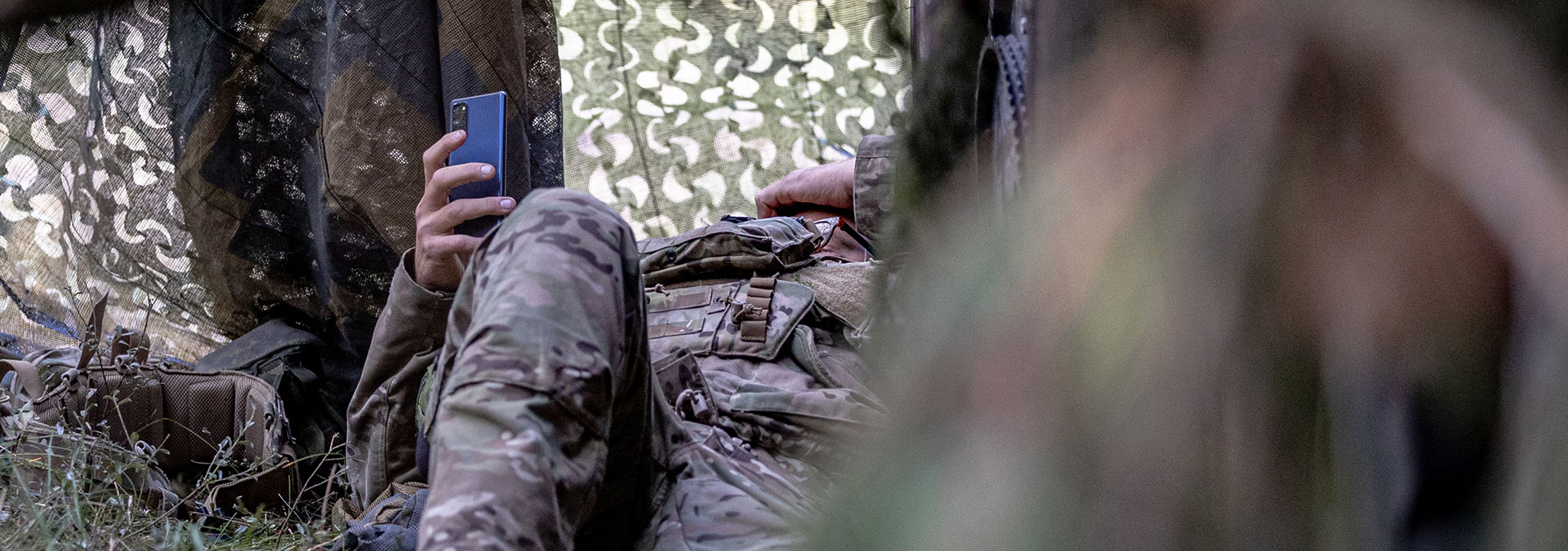 En soldat i uniform ligger ned i et aflukke skabt af sløringsnet med armen hævet højt og en mobiltelefon i hånden.