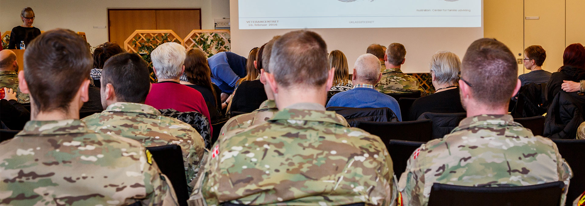 I en større sal sidder uniformerede soldater med deres pårørende i civilt tøj . De sidder med ryggen til på stolerækker foran et powerpointshow og lytter på et oplæg fra en kvinde.