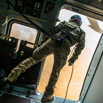 En af besætningen står i helikopterens åbning og ser ud på brun støv, som helikopteren hvirvler op fra jorden