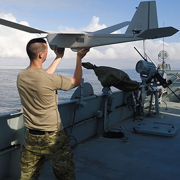 Mand står i uniform på skibsdæk og holder en drone i hånden.