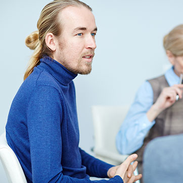 Langhåret tynd mand med håret i en elastik sidder på en stol i et lokale, hvor man kan ane en kvinde. Han vender ansigtet mod en anden og har hænderne i skøddet.