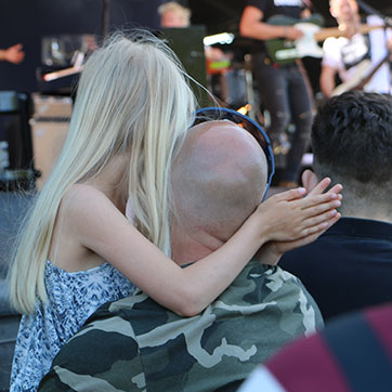 Med scenen i baggrunden ses ryggene af koncertpublikum. En skaldet mand i camouflagefarvet t-shirt bærer sin datter, så hun kan se scenen. Hun er iført høreværn og klapper bag hans nakke.