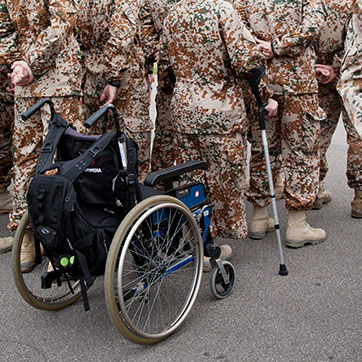 Tom kørestol bagerst i en række af soldater. Foran kørestolen står en uniformeret soldat på krykker.