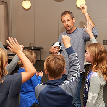 Mand med skæg står foran en gruppe af børn med en bold hvilende på sin ene underarm, mens han rækker den anden hånd i vejret. Børnene kopierer håndsoprækningen.