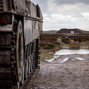 En kampvogn af mærket Leopard sigter mod nogle skiver på en forhøjning længere fremme i et kuperet terræn, primært dækket af lyng. Himlen er gråmeleret, og der er vandpytter i gruset foran kampvognen.