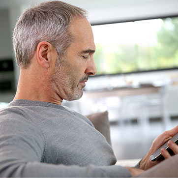 Ældre mand med kort, gråt hår og grånet skæg i en sofa med sin smartphone i hånden. Hjemmet er lyst med et stort vindue for enden.