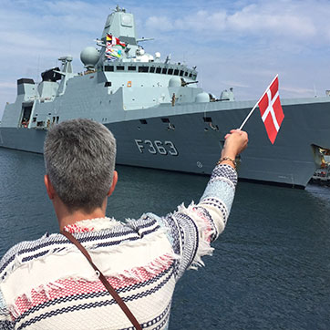 Karseklippet kvinde står i striktrøje på havnekaj og vifter med et plastikflag mod gråt skib, korvetten Niels Juel.