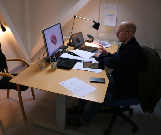 Nærbillede af en hånd, der holder en kuglepen, imens pegefingeren trykker på en tablet, der ligger på et bord. Der ligger papirer ved siden af tabletten.