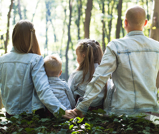 En familie bestående af fra venstre mod højre, en kvinde, to mindre børn og en mand sidder tæt sammen på en bund af efeu i en skov, hvor solens stråler lyser igennem trækronerne.
