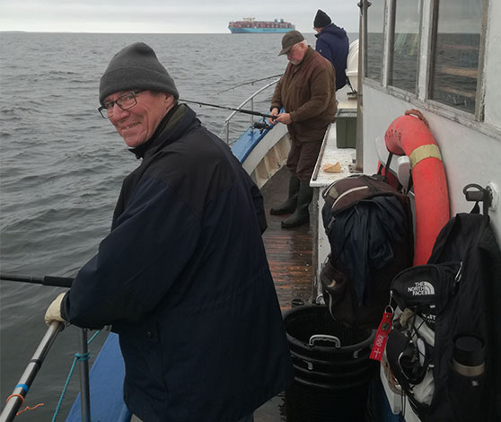 Tre mænd med varm tøj og huer er i gang med fiskestænger på en mindre fiskekutters dæk. Der ligger spande og udstyr mellem dem. Manden nærmest billedet smile.