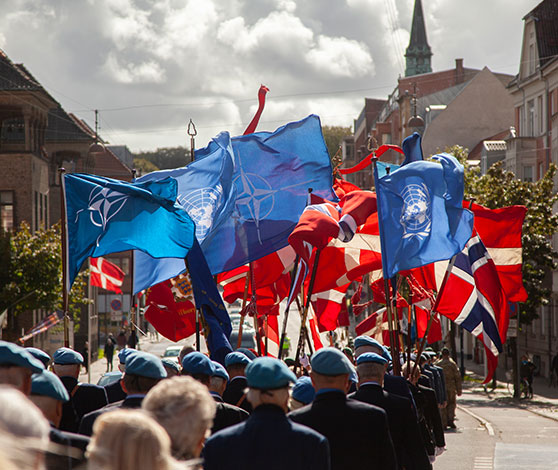 Flagparade med adskillige danske faner og enkelte blå NATO-faner. Fanebærerne ses gå gennem Aalborg midtby.
