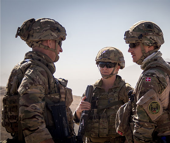 To soldater i fuld udrustning med veste, hjelme og geværer lytter til deres overordnede.