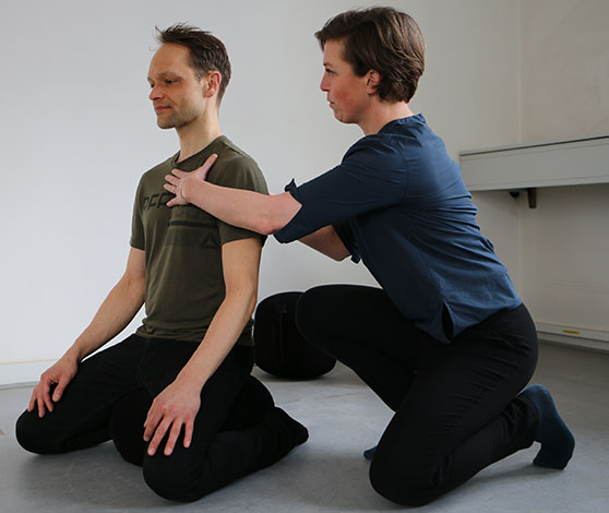 Mand sidder på knæ på gulvet med lukkede øjne, mens kvinde knæler og instruerer ham ved at lægge en hånd på hans brystkasse og ryg.