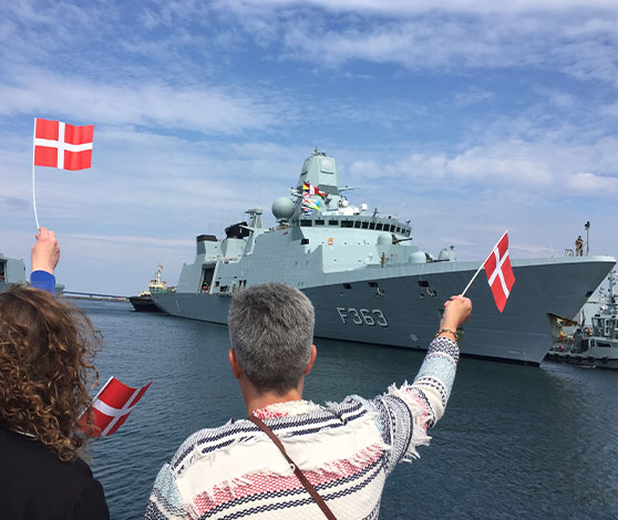 Folk på kajkanten vinker med flag ud mod fregatten Niels Juel, der er på vej i havn.