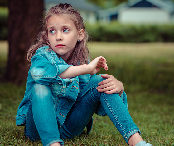 En pige, der nok er mellem 8-12 år, sidder på en græsplæne med armene hvilende på knæene. Hun ser sig let til siden og har en rynke i panden, midt mellem øjenbrynene.