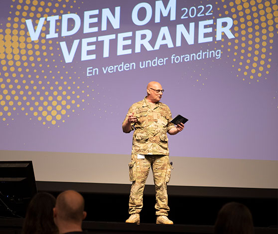 En skaldet mand i uniform står alene på en stor scene og taler med en blå baggrund, hvorpå der står Viden om veteraner.